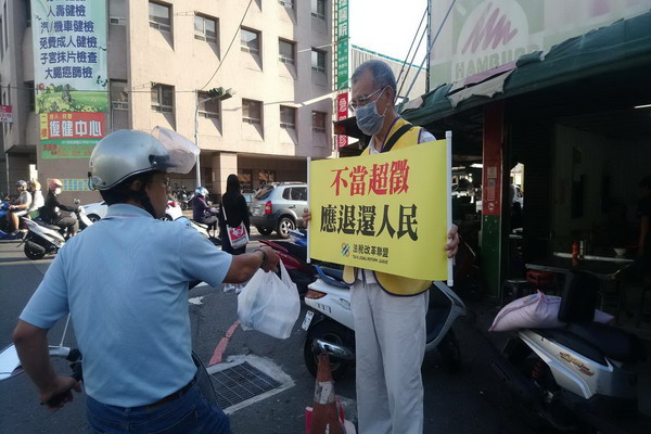 年近80歲的魏姓志工在中區國稅局大屯稽徵所前舉牌街宣時,男士手提四瓶冰涼的礦泉水慰勞志工的辛勞。